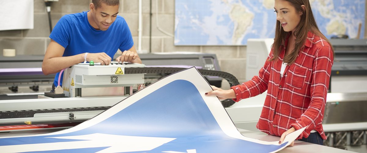 A young woman is working on a digital printing machine . she is printing out a large one way sign . A colleague is resetting the printer heads . ** background map has been modified**